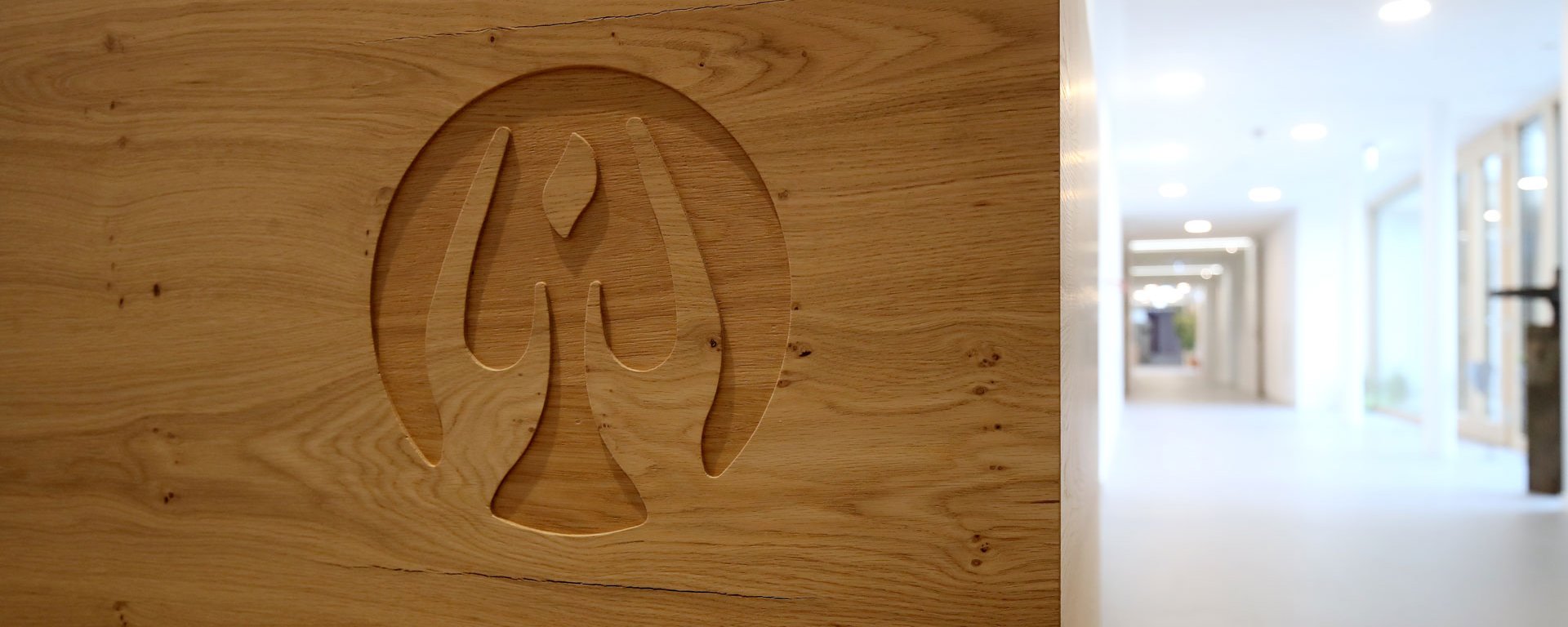 In Holz geschnitztes Logo des AllgäuHospiz mit Blick in die Tiefe der Gänge des AllgäuHospiz